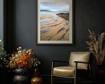 May include: A framed landscape photograph depicting a sandy beach with dark, rippled patterns leading toward the ocean, rocky formations on the right, and a cloudy sky, displayed on a dark wall above a leather armchair and dried flowers.