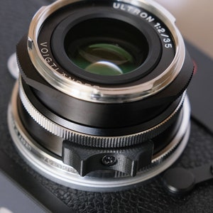 Peut inclure: Un objectif d'appareil photo noir et argent avec le texte "ULTRON 1:2/35" et "VOIGTLANDER" imprimé sur le côté.