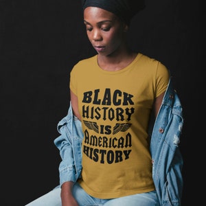 Puede incluir: Una camiseta amarilla mostaza con texto negro que dice "Black History is American History" con alas a cada lado de la palabra "is".