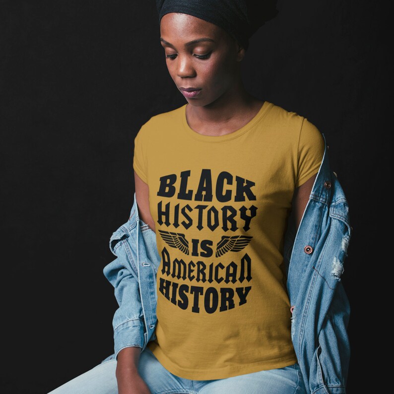 Puede incluir: Una camiseta amarilla mostaza con texto negro que dice "Black History is American History" con alas a cada lado de la palabra "is".