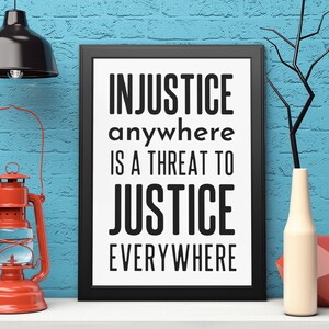 Puede incluir: Impresi&oacute;n enmarcada en negro con el texto "INJUSTICE anywhere IS A THREAT TO JUSTICE EVERYWHERE" sobre fondo blanco. La impresi&oacute;n est&aacute; en un estante blanco con una linterna roja y un jarr&oacute;n blanco con una rama.
