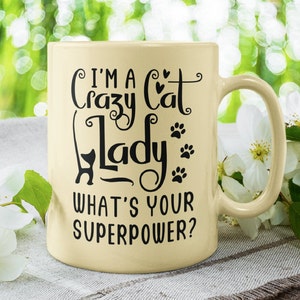 May include: A cream-colored ceramic mug with a black design that reads "I'm a crazy cat lady. What's your superpower?" The mug has a black cat silhouette and paw prints.