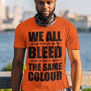Puede incluir: Camiseta naranja con el texto "WE ALL BLEED THE SAME COLOUR" en negro.