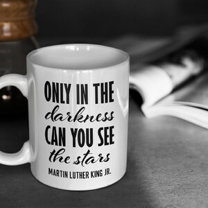 Puede incluir: Taza de caf&eacute; de cer&aacute;mica blanca con una cita en negro que dice "Only in the darkness can you see the stars - Martin Luther King Jr."