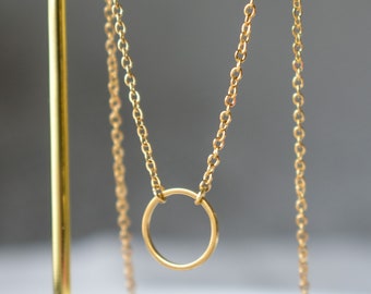 Könnte beinhalten: Eine zarte, goldfarbene Halskette mit einem kleinen, offenen Kreisanhänger. Der Anhänger hängt an einer feinen Kette und erzeugt ein minimalistisches Design. Die Halskette wird auf einem goldfarbenen Ständer vor einem weichen, verschwommenen Hintergrund präsentiert.