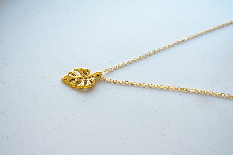 Peut inclure: Un collier en or avec une cha&icirc;ne d&eacute;licate et un pendentif en forme de feuille. Le pendentif est une d&eacute;coupe d&eacute;taill&eacute;e d'une feuille de monstera.