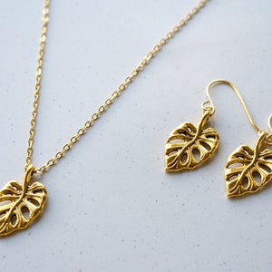 Peut inclure: Un ensemble de collier et boucles d'oreilles en or avec un motif d&eacute;licat de feuille de monstera. Le collier a une cha&icirc;ne simple et les boucles d'oreilles sont de style pendantes.