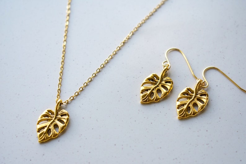 Peut inclure: Un ensemble de collier et boucles d'oreilles en or avec un motif d&eacute;licat de feuille de monstera. Le collier a une cha&icirc;ne simple et les boucles d'oreilles sont de style pendantes.