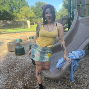 May include: A woman wearing a yellow tank top, denim mini-skirt with patches, and black boots. She is smiling and leaning against a playground slide, with a denim jacket nearby. A red and purple bandana is on her head.