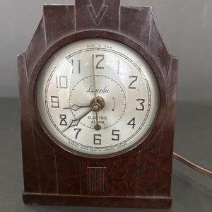 May include: A vintage brown Bakelite electric alarm clock with a white face and black hands. The clock face reads "Lincoln Electric Alarm" and "Electric Clock Corp of America Chicago".