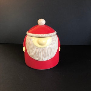 May include: A red ceramic cookie jar shaped like a Santa Claus with a white beard and a yellow nose. The jar has a lid with a white trim.