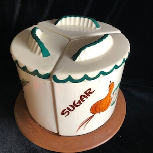 May include: A white ceramic sugar dispenser with a green trim and a brown wooden base. The dispenser has a pumpkin design and the word "SUGAR" written on it.