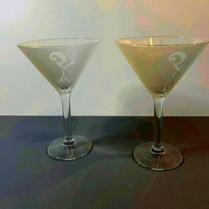 May include: Two clear martini glasses with a frosted finish. Each glass has a long stem and a wide, triangular bowl. A decorative design is etched on the front of each glass. The glasses are set against a dark surface.