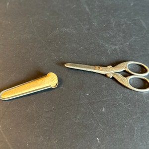 May include: A silver pair of small scissors with an ornate design and a matching beige leather case.