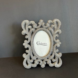 Puede incluir: Un marco de fotos plateado con un diseño ornamentado. El marco tiene una abertura ovalada y la palabra "Chehoma" está impresa en negro dentro del marco.
