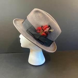 Pode incluir: Um chapéu fedora cinza com uma faixa de fita de gorgorão preta e um detalhe decorativo de penas. O chapéu é exibido em uma cabeça de manequim branca contra um fundo escuro. O detalhe de penas apresenta cores vermelhas, pretas e douradas.