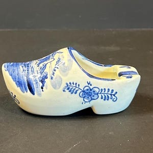 May include: A ceramic clog-shaped ashtray, painted in white and blue. The ashtray features a floral design and landscape details in blue. The interior is a light yellow color. A decorative item.