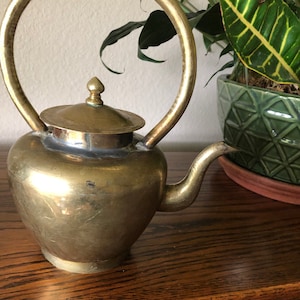 Può includere: Una teiera in ottone con un design di manico unico. La teiera ha un corpo rotondo e un coperchio con un piccolo pomo in cima. La teiera è appoggiata su una superficie di legno.