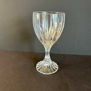 May include: A clear glass wine goblet with a tall, slender stem and a wide, fluted bowl. The goblet has a decorative, ribbed pattern.