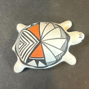 May include: A ceramic turtle figurine with a cream-colored body and a shell decorated with geometric patterns in black, white, and orange. The turtle has four flipper-like feet and a rounded head.
