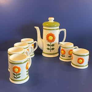 Puede incluir: Juego de café de cerámica vintage con una tetera blanca con tapa verde y diseño de girasol naranja. Incluye tazas y una jarra de crema a juego. El juego tiene una estética retro con detalles florales.