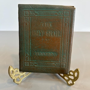 May include: A small, green leather-bound book titled "The Holy Grail" by Tennyson, displayed on a gold metal stand.