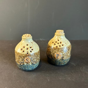 Puede incluir: Un juego de dos saleros y pimenteros de cerámica con tapas de corcho. Los saleros son marrones, beige y azules con un esmalte moteado. Los saleros tienen agujeros para dispensar sal y pimienta. Los saleros están etiquetados con "S" y "P".