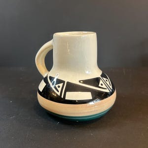 Pode incluir: Uma pequena caneca de cerâmica com uma alça. A caneca tem um topo creme e uma base estampada com faixas pretas, brancas, bege e verdes. O design apresenta formas geométricas. A caneca é provavelmente feita à mão.
