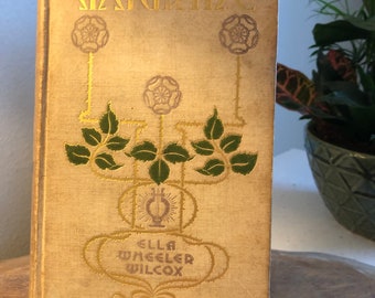 Könnte beinhalten: Ein Vintage-Buch mit dem Titel "Dainty" von Ella Wheeler Wilcox. Der Einband zeigt ein Blattgold-Design mit Rosen und Blättern auf cremefarbenem Hintergrund.