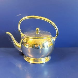 Może przedstawiać: Biały ceramiczny kubek z czarnym uchwytem i napisem "But First, Coffee" pogrubioną, czarną czcionką. Kubek jest ustawiony na rozmytym tle, sugerującym przytulne otoczenie.