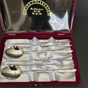 Puede incluir: Un juego de palillos y cucharas de plata en una caja de terciopelo rojo con forro de satén blanco. La caja tiene una etiqueta dorada que dice "Living Collection Best Design for your Good Life Your Eyes Only".