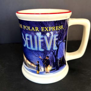 Op de afbeelding: Een witte keramische mok met een rode rand en een handvat, voorzien van een afbeelding uit The Polar Express. Het ontwerp toont een besneeuwd nachttafereel met een conducteur en een kind, de tekst THE POLAR EXPRESS en het woord BELIEVE.