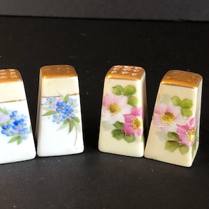Op de afbeelding: Set van vier vintage zout- en peperstrooiers met bloemmotieven. Twee strooiers hebben roze rozen en groene bladeren, terwijl de andere twee blauwe vergeet-me-nietjes en groene bladeren hebben. Elke strooier heeft een gouden rand aan de bovenkant.