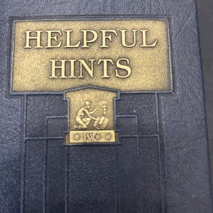 Può includere: Un libro rilegato in pelle blu con lettere dorate che recitano "HELPFUL HINTS". La copertina presenta un disegno dorato di una persona che tiene una torcia e un libro.