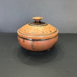 Op de afbeelding: Een bruine keramische kom met deksel, een zwarte rand en een getextureerd ontwerp op het deksel. De kom heeft een glanzende afwerking.