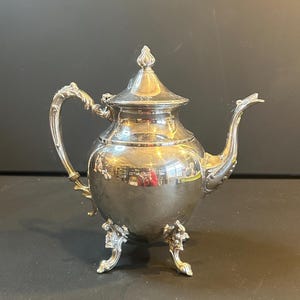 Puede incluir: Una tetera plateada con detalles ornamentados. La tetera tiene un asa curvada, un pico y una tapa con un adorno decorativo. Se apoya sobre tres patas decorativas. La superficie pulida refleja el entorno.