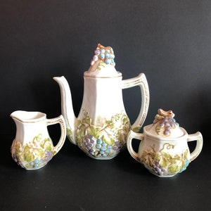 Op de afbeelding: Een wit porseleinen theeservies met een druivenrankenontwerp. De theepot, roomkan en suikerpot zijn versierd met groene bladeren en paarse druiven.