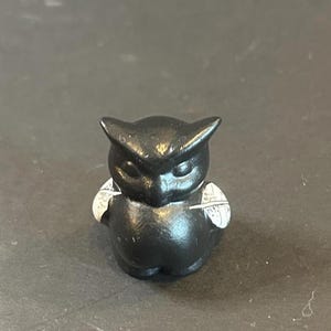 May include: A small, black owl figurine with white accents on its wings. The owl has a rounded body, pointed ears, and closed eyes, giving it a serene appearance. The figurine is likely made of a solid material.