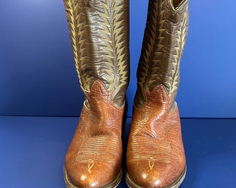Può includere: Un paio di stivali da cowboy in pelle marrone. Gli stivali hanno la punta affusolata e un gambale alto con cuciture decorative. La tomaia è di una tonalità marrone più chiara, mentre il gambale è marrone più scuro con un design dorato. Gli stivali sono su sfondo blu.