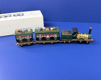 May include: A miniature toy train set featuring a green steam locomotive numbered 25, followed by two green passenger cars with gold trim and luggage racks on top. The cars feature pink curtains and small windows. A white box is positioned behind the train.