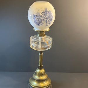 May include: An antique oil lamp with a round, frosted glass shade featuring a blue and white landscape scene. The lamp has a clear glass reservoir, brass accents, and a sturdy base. A vintage decorative piece.