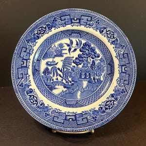 Pode incluir: Prato de cerâmica azul e branco com design Willow. O prato tem uma borda detalhada e uma cena central que retrata uma paisagem com uma ponte, árvores e pássaros. O prato é redondo e tem uma estética clássica e vintage.