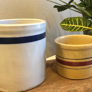 May include: Two antique stoneware crocks. The larger crock is off-white with a blue band. The smaller crock is tan with red and blue bands. Both have a cylindrical shape and are likely vintage kitchenware.