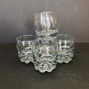 May include: Four clear glass tumblers with a raised bottom and a rounded shape. Each tumbler has the text "McCormick" etched into the glass.