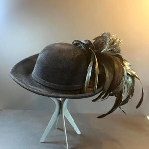 Puede incluir: Un sombrero negro con una copa redondeada y un ala ancha, adornado con un grupo de plumas negras. El sombrero se exhibe sobre un pequeño soporte de tres patas de color gris claro, sobre un fondo neutro.
