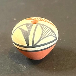 May include: A hand-painted, spherical ceramic bead with a hole. The bead features a cream-colored background with black and gray geometric designs, a red base, and a small red accent on top. The bead is likely for jewelry making.