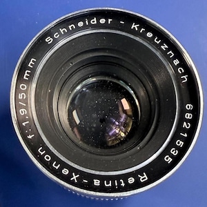 May include: A black and silver Schneider-Kreuznach Retina-Xenon f:1.9/50 mm camera lens. The front element is circular with visible glass reflections. The outer ring features white engraved text including the brand, model, and serial number 6821535.