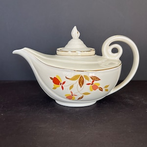 May include: A white teapot with a gold trim and a floral pattern in shades of orange, yellow, and brown. The teapot has a unique, curved spout and a lid with a knob in the shape of a flame.
