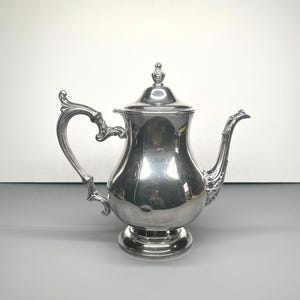 Peut inclure: Une théière argentée avec une anse et un bec courbés. La théière a un couvercle avec un bouton décoratif et repose sur une base ronde. La théière est probablement en métal et reflète l'environnement.