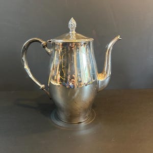 May include: A silver-colored teapot with a curved handle and a spout. The teapot has a lid with a decorative finial. The pot is sitting on a dark surface.